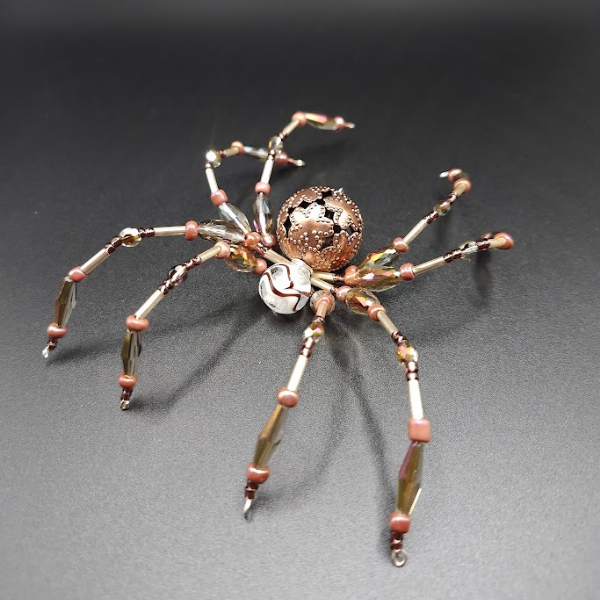 Beaded spider-shaped jewelry piece with a spherical, floral-patterned body on a dark background.