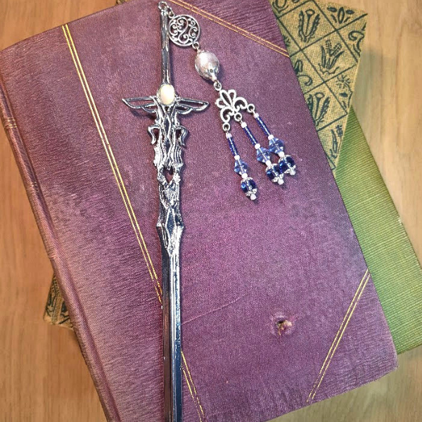A silver sword bookmark and a blue beaded charm resting on a stack of three closed books, with the top book being purple with gold markings, and the other books green and dark green, on a wooden surface.