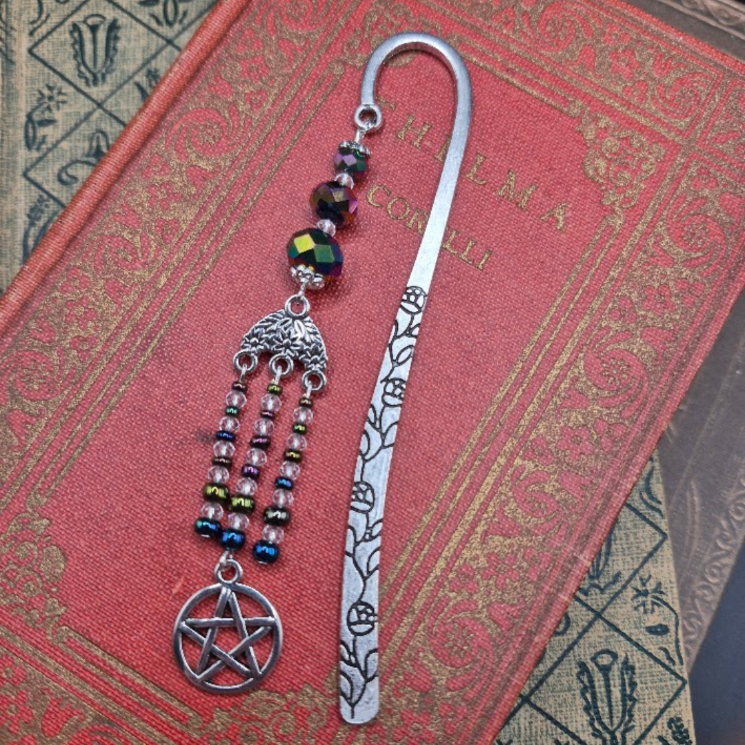 A decorative bookmark with a pentagram pendant and beaded dangles, placed on a red book. The bookmark has a silver-colored handle with floral cutouts and multicolored beads.