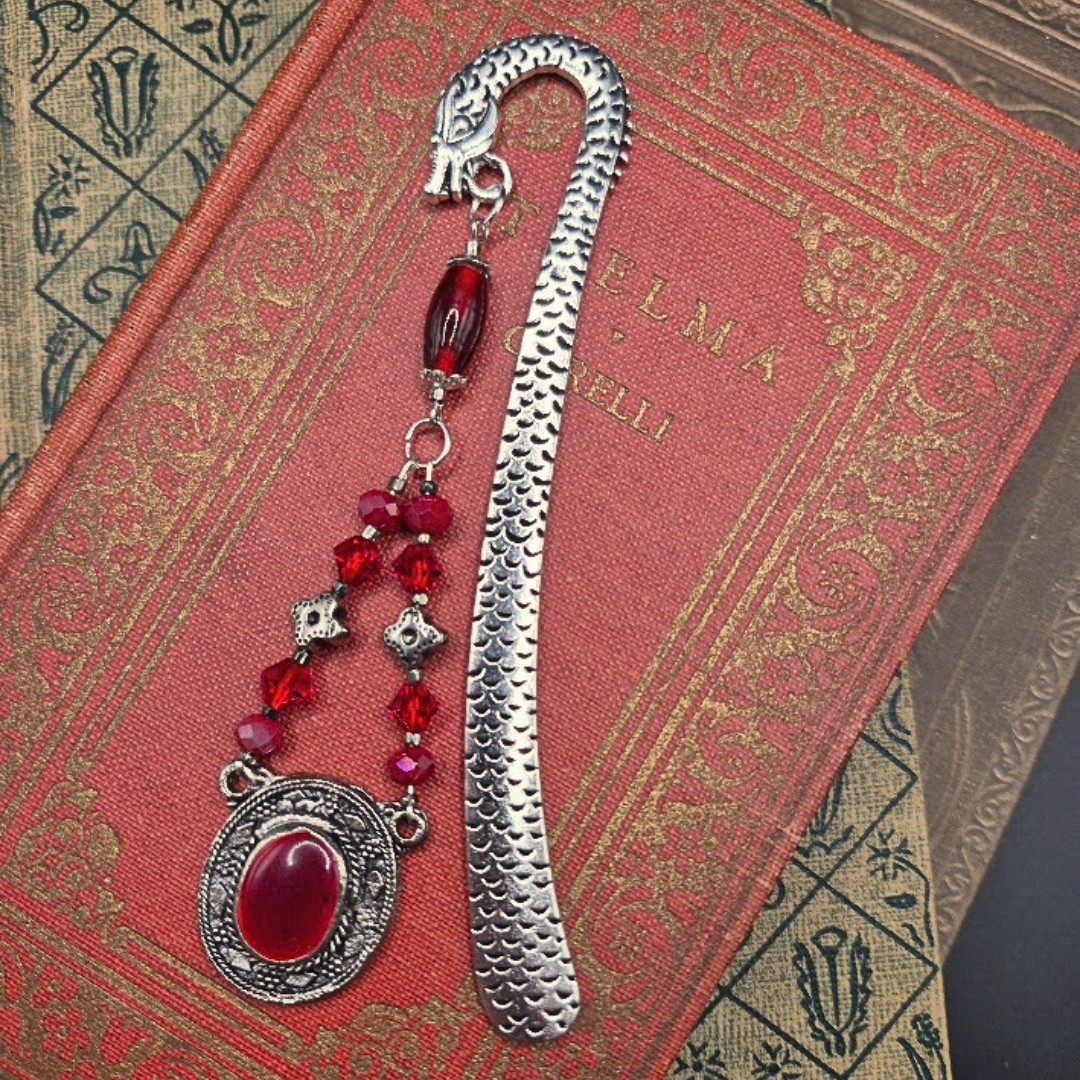 A red book with a metal bookmark featuring red beads and silver decorative elements resting on top.