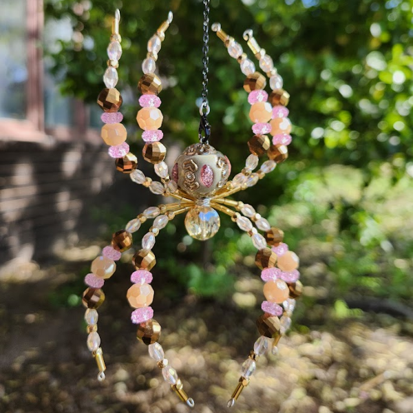 Beaded spider ornament with pink, gold, and clear beads, hanging outdoors with green trees in the background.