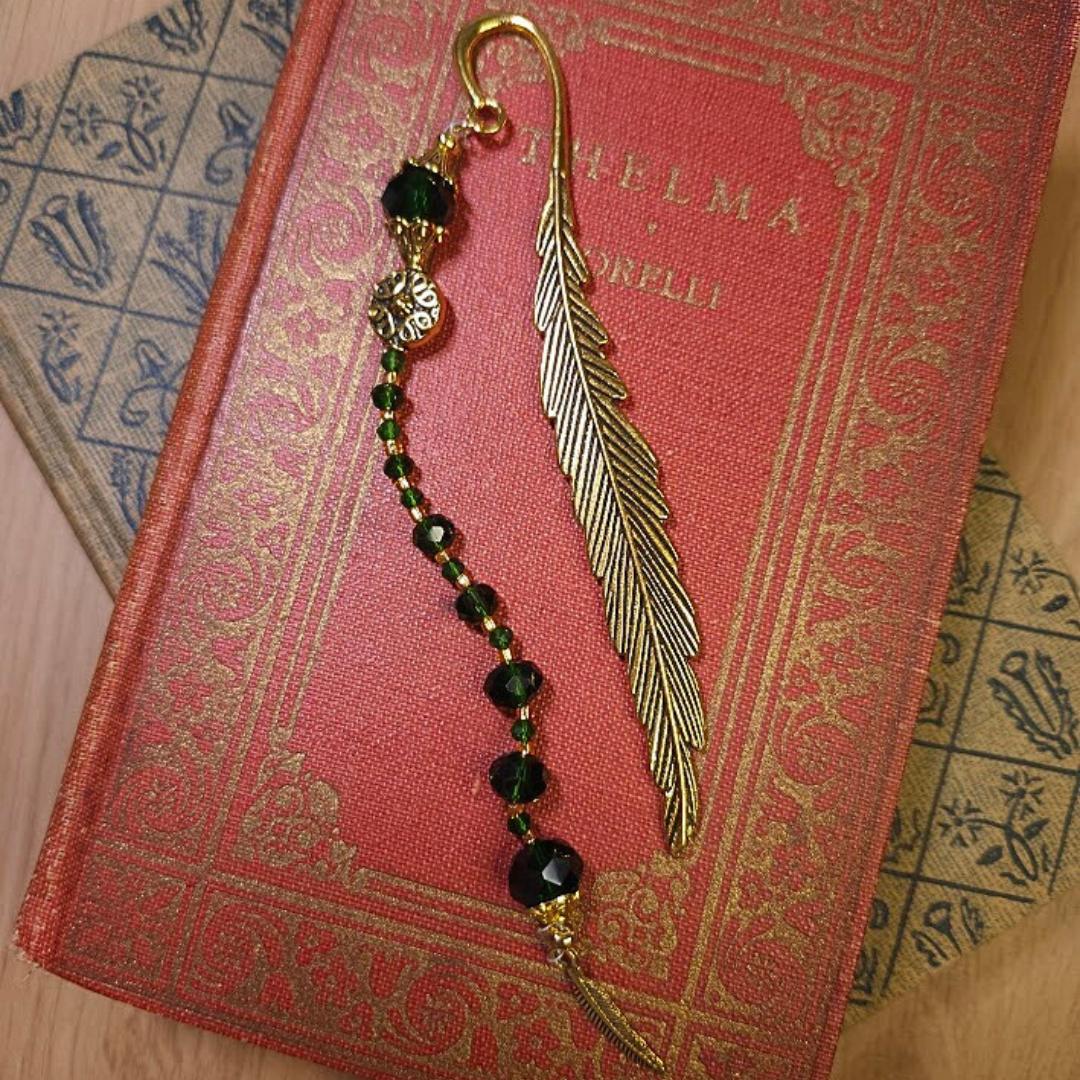 A gold feather-shaped bookmark with a beaded charm, placed on a red book titled 'CAMILLA LACKS'. The beads are black, green, and gold, and there is additional decorative gold detailing on the bookmark.