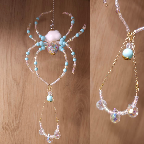 A handcrafted spider-shaped ornament made of beads, including a large central iridescent bead, blue beads, and clear beads, with a chain and bead charm hanging below.