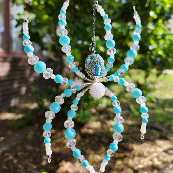 Colorful beaded spiderwith blue, white, and clear beads, hanging outdoors with greenery in the background.