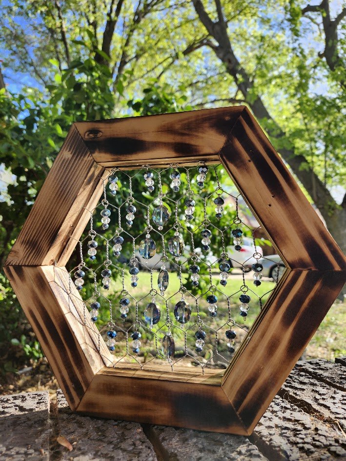 A wooden hexagonal suncatcher with hanging glass beads and crystals, placed outdoors on a piece of wood with green trees and blue sky in the background.