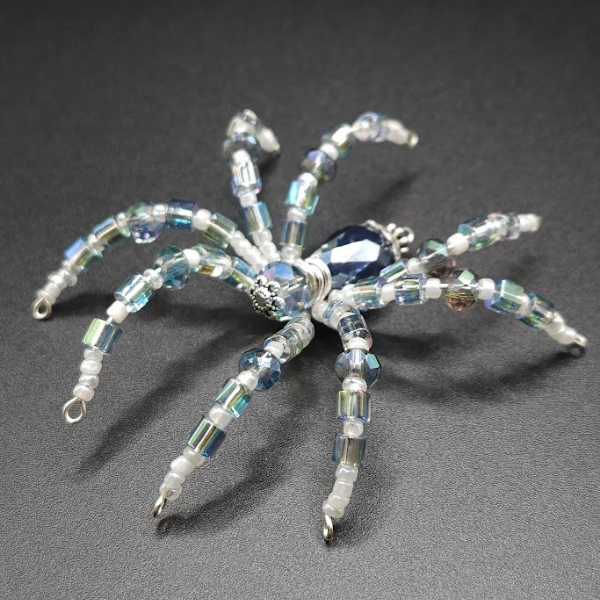 Beaded spider sculpture made of iridescent beads with a large blue center bead, set on a black background.