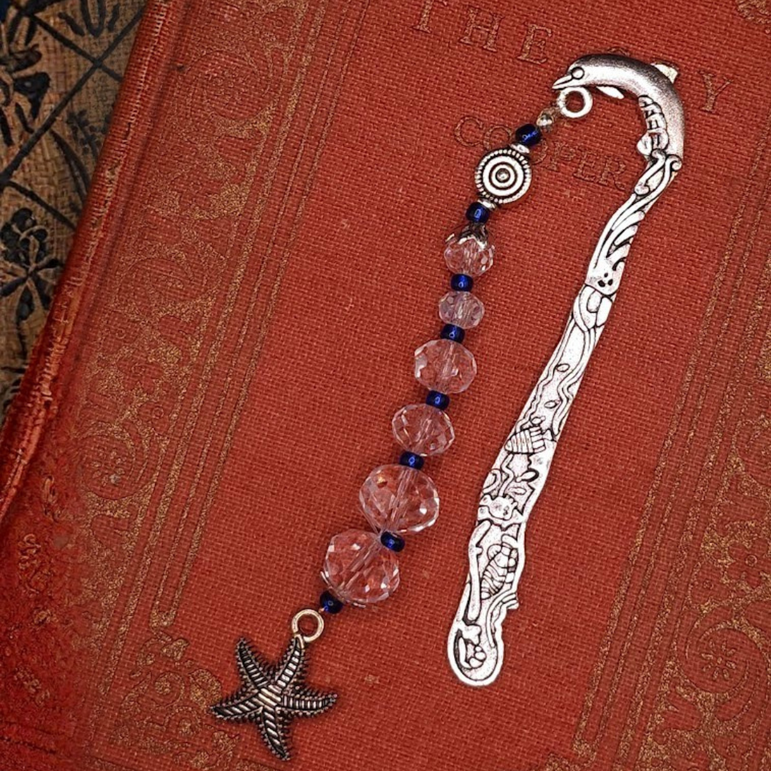 A decorative metal bookmark with beads and a starfish charm on a red textured book cover.