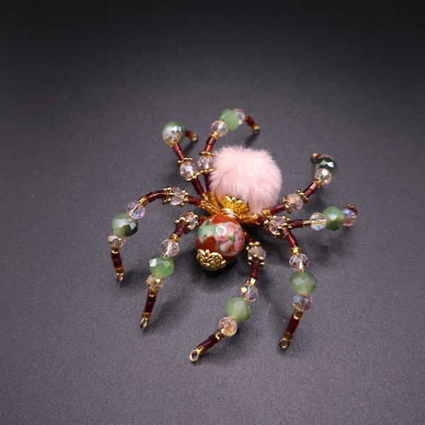 Decorative beaded spider jewelry with pink fuzzy body on a dark background.
