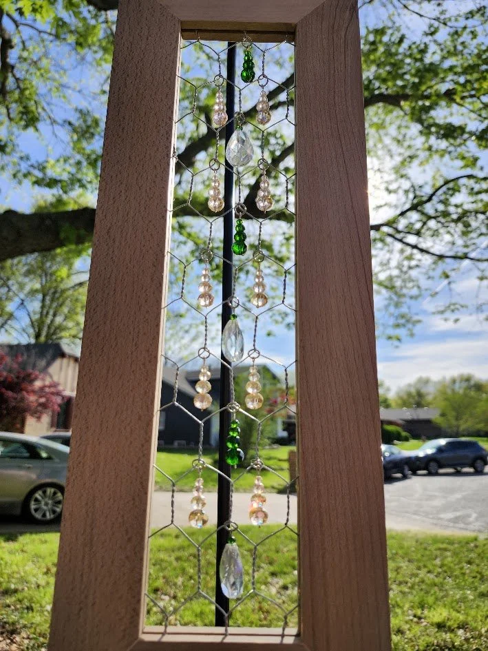 A wooden framed suncatcher with decorative hanging beads and crystals, with a background of trees, blue sky, and residential houses.