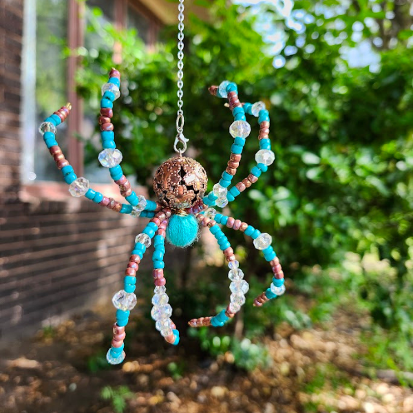 Beaded spider ornament hanging outdoors amid greenery and a brick wall.