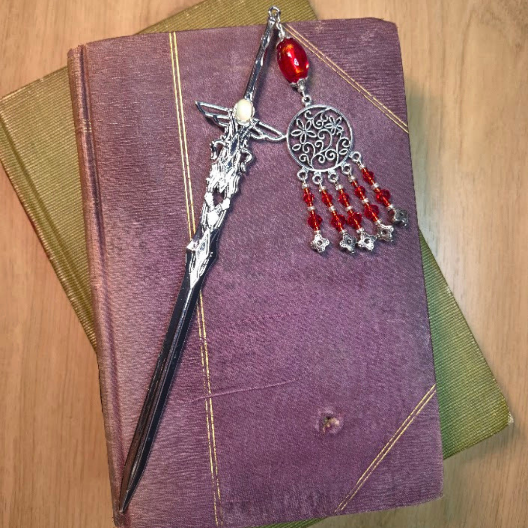 A purple book with a green spine, a silver sword bookmark with a decorative charm with red beads and a round centerpiece with an intricate design placed on top of it.