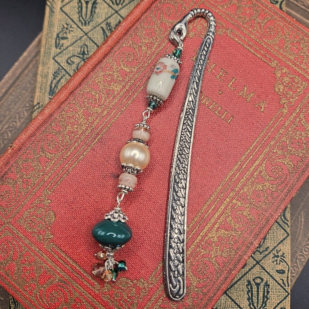 A decorative bookmark with a dragon-shaped metal top, a floral ceramic bead, a pearl, and a teal bead hangs over a red, gold-embossed book, resting on another vintage book with intricate patterns.