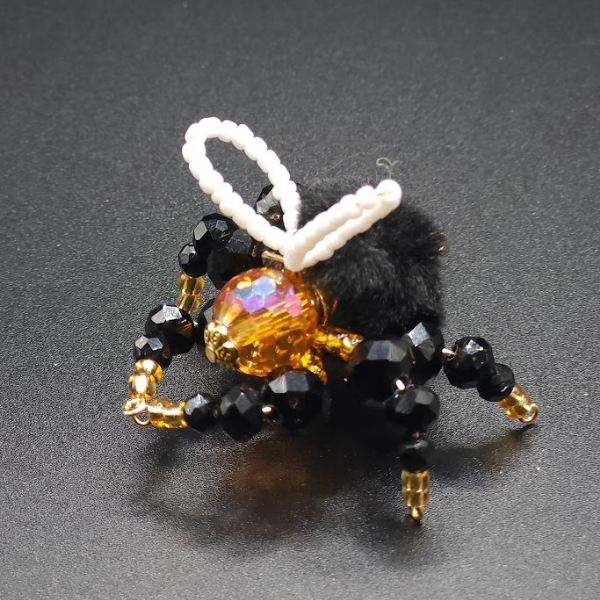 Beaded decorative bee with black and gold beads, a shimmering gold and orange bead body, a black fuzzy thorax, white beaded wings, and a fuzzy black tail on a black background.