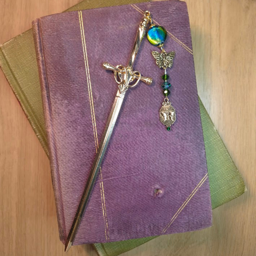 An ornate silver sword bookmark with a colorful decorative chain with beads and charms resting on two stacked closed books, one purple and one green, on a wooden surface.
