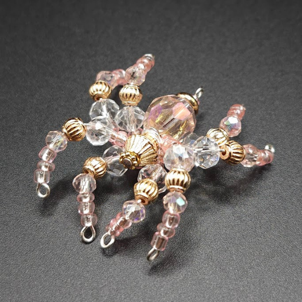 Close-up of a decorative spider-shaped jewelry piece with pink, clear, and gold beads on a dark background.
