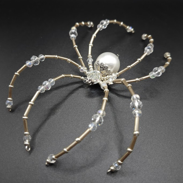 Beaded spider with metallic and beaded legs and a large pearl body, set on a black background.