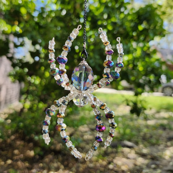 A beaded spider ornament with colorful, iridescent beads, hanging outdoors against a green, leafy background.