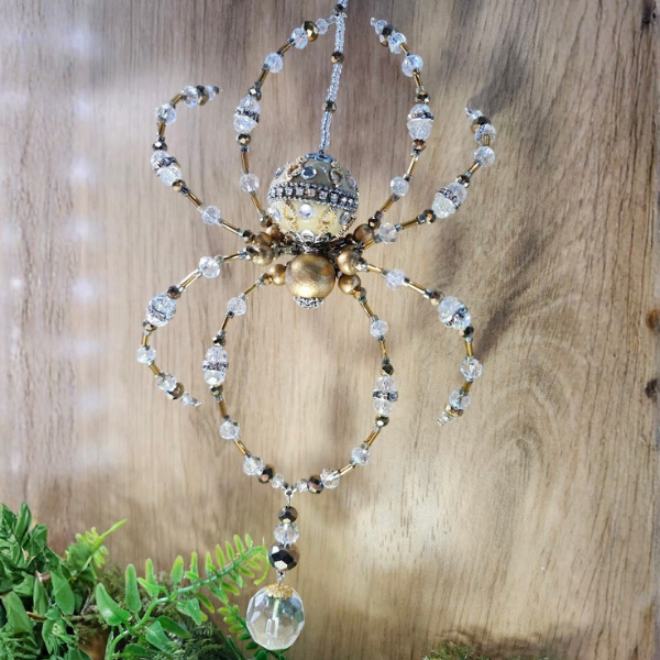 Decorative spider ornament made with beads and crystals hanging from a wooden surface, with green plants at the bottom.