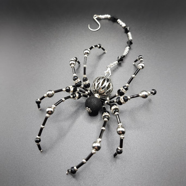 Jewelry piece in the shape of a spider made of black and silver beads with a spherical pendant, displayed on a dark gray surface.