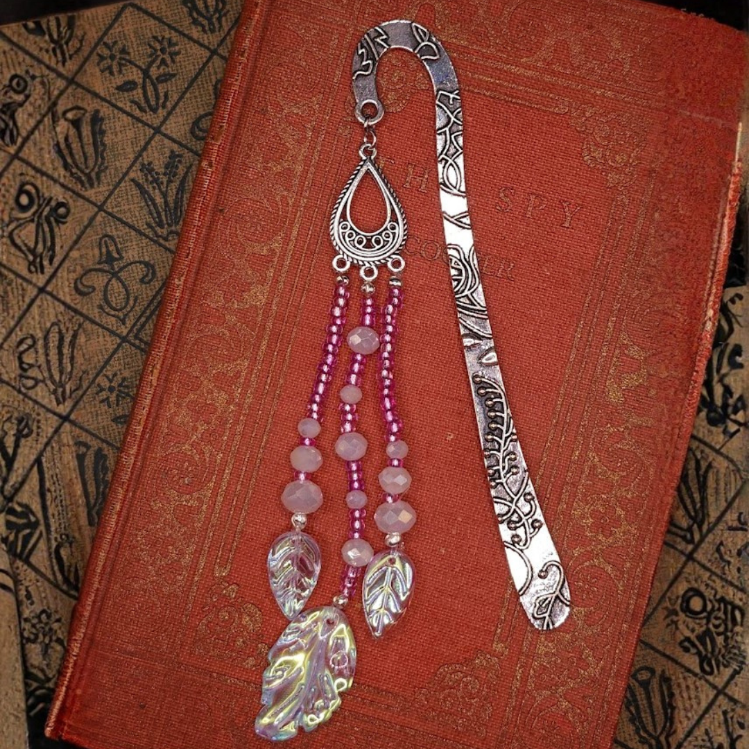 A decorative silver bookmark with intricate patterns and dangling pink beads, resting on a red vintage book with a textured cover.