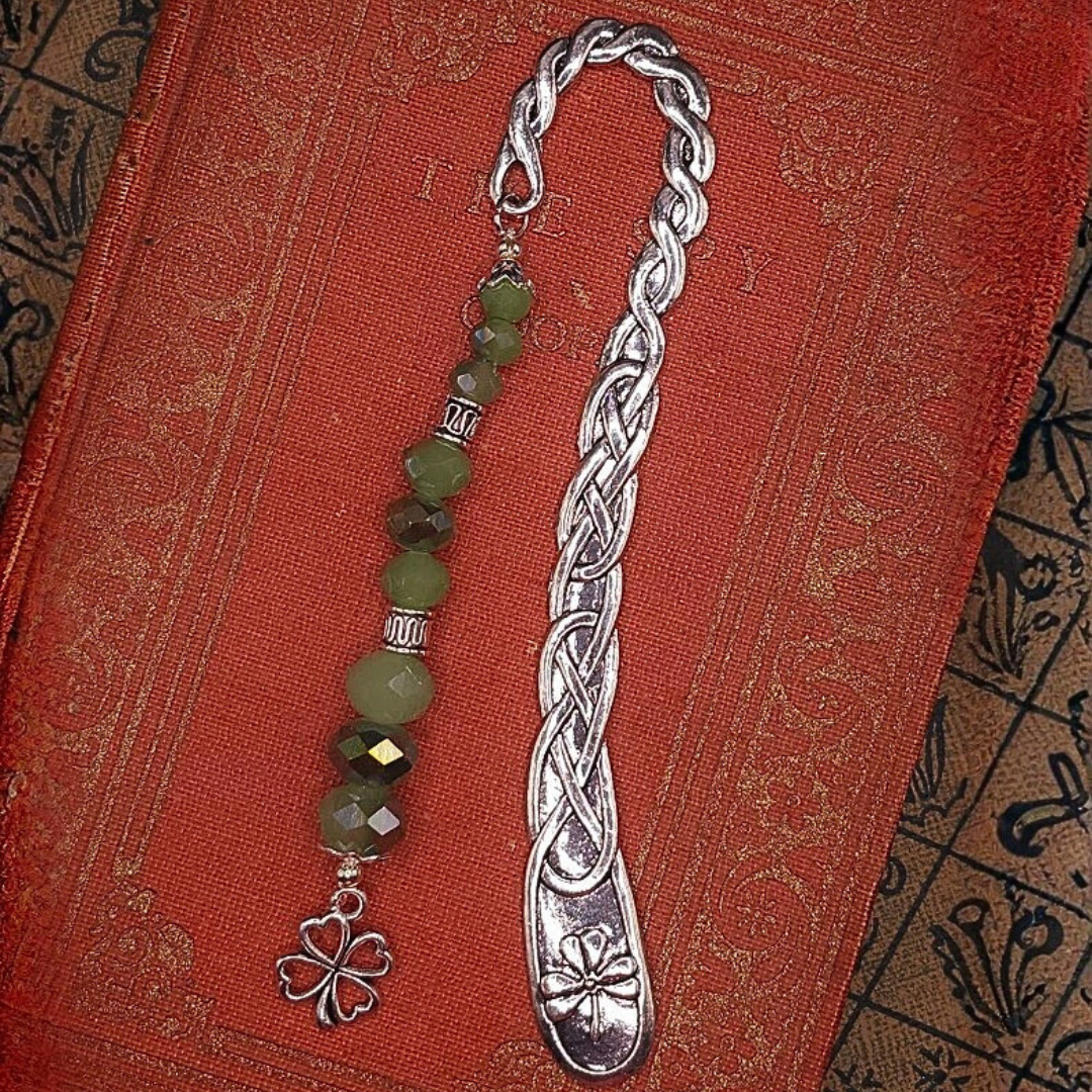 A silver hook bookmark with an intricate braided design and a beaded charm with green beads and a four-leaf clover pendant, placed on a red textured book cover.