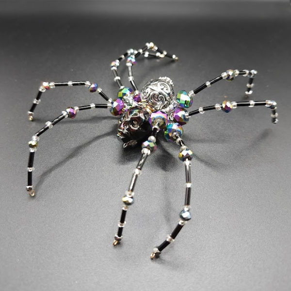 A spider piece made of black and iridescent beads with metallic accents on a black surface.