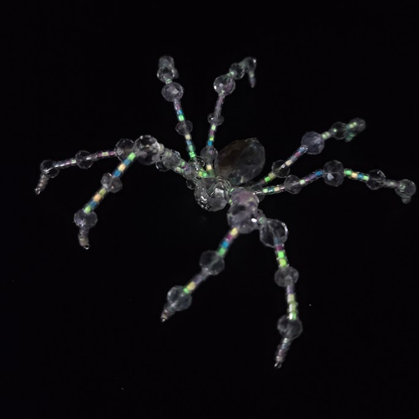 A spider-shaped ornament made of clear, iridescent beads and tiny metallic beads, set against a black background.