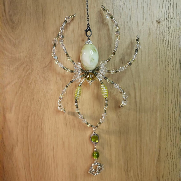 Decorative spider-shaped ornament featuring a large central pearl, beaded legs, and dangling green and silver elements, hanging against a wooden background.