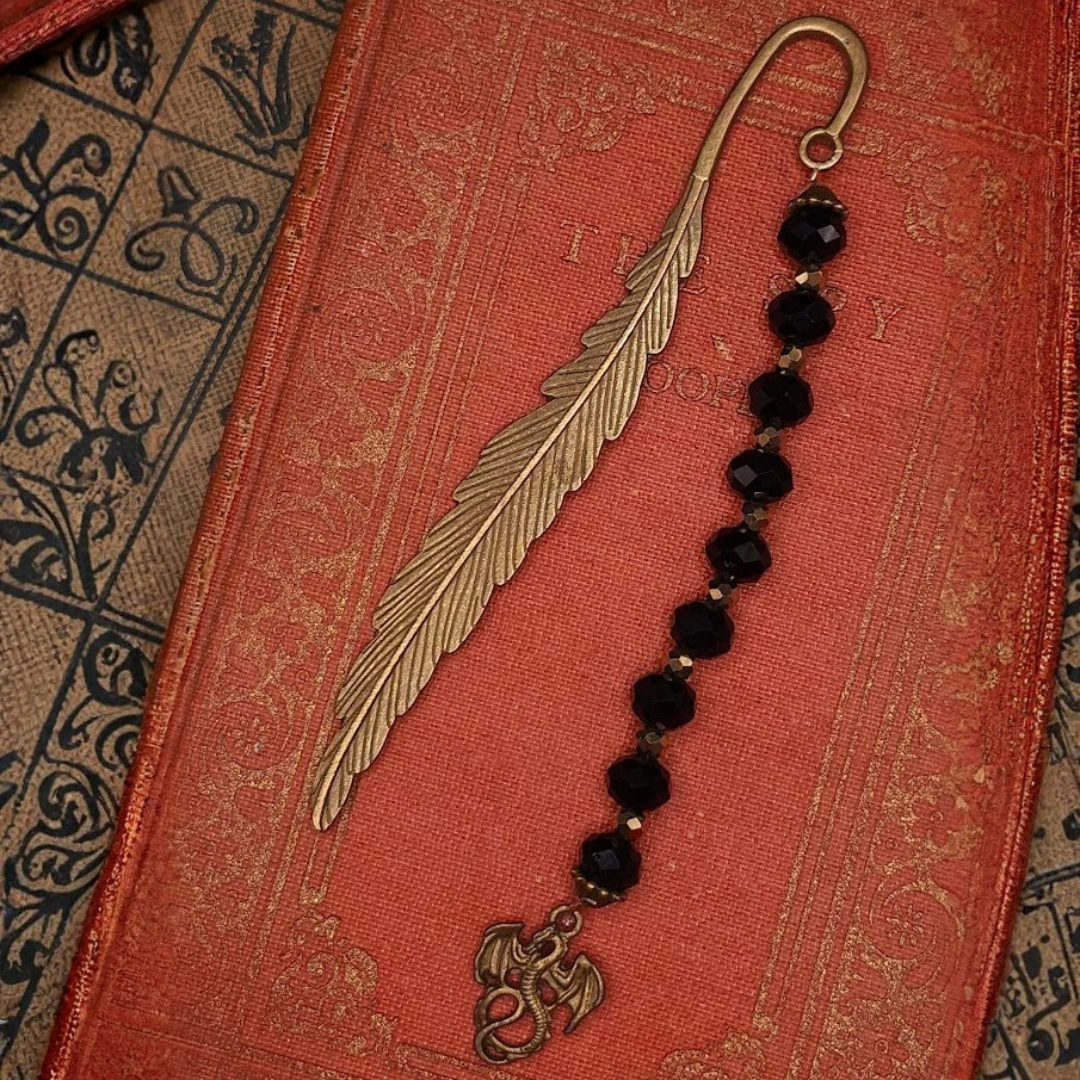 A vintage gold feather bookmark and a black beaded bracelet with a dragon charm on a red decorative book cover.