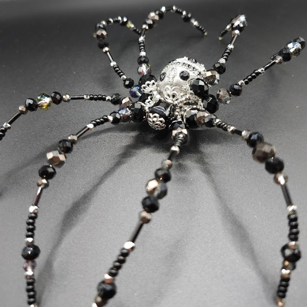 A beaded black spider jewelry piece with a silver decorative head and bead legs on a black background.