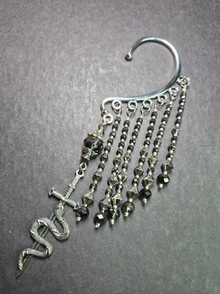 A decorative ear cuff with beads, black beads, and charms including a snake and a sword or dagger.