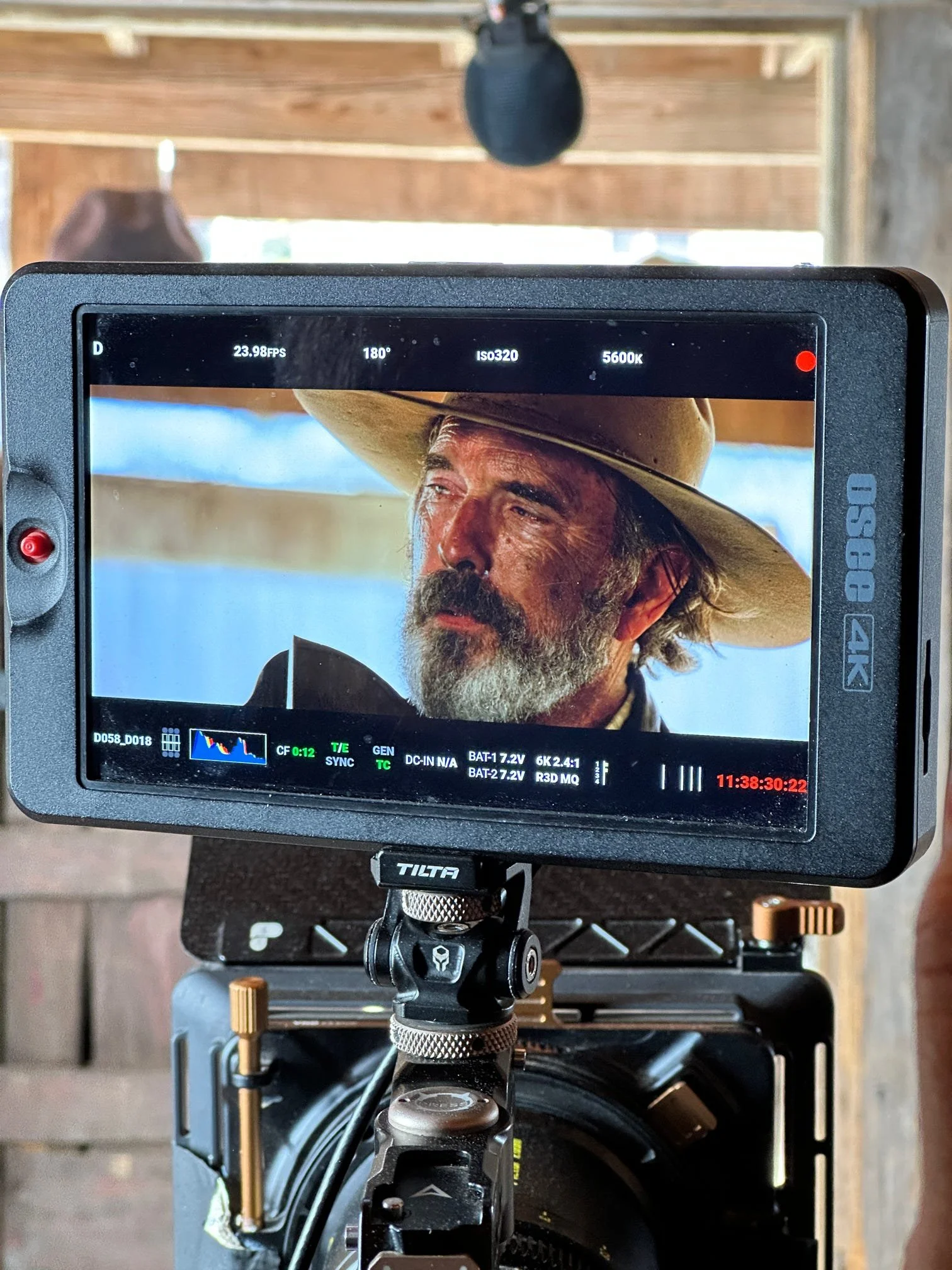 Close-up of a camera monitor showing a man with a beard and mustache, wearing a cowboy hat, with other equipment and a wheel below it.