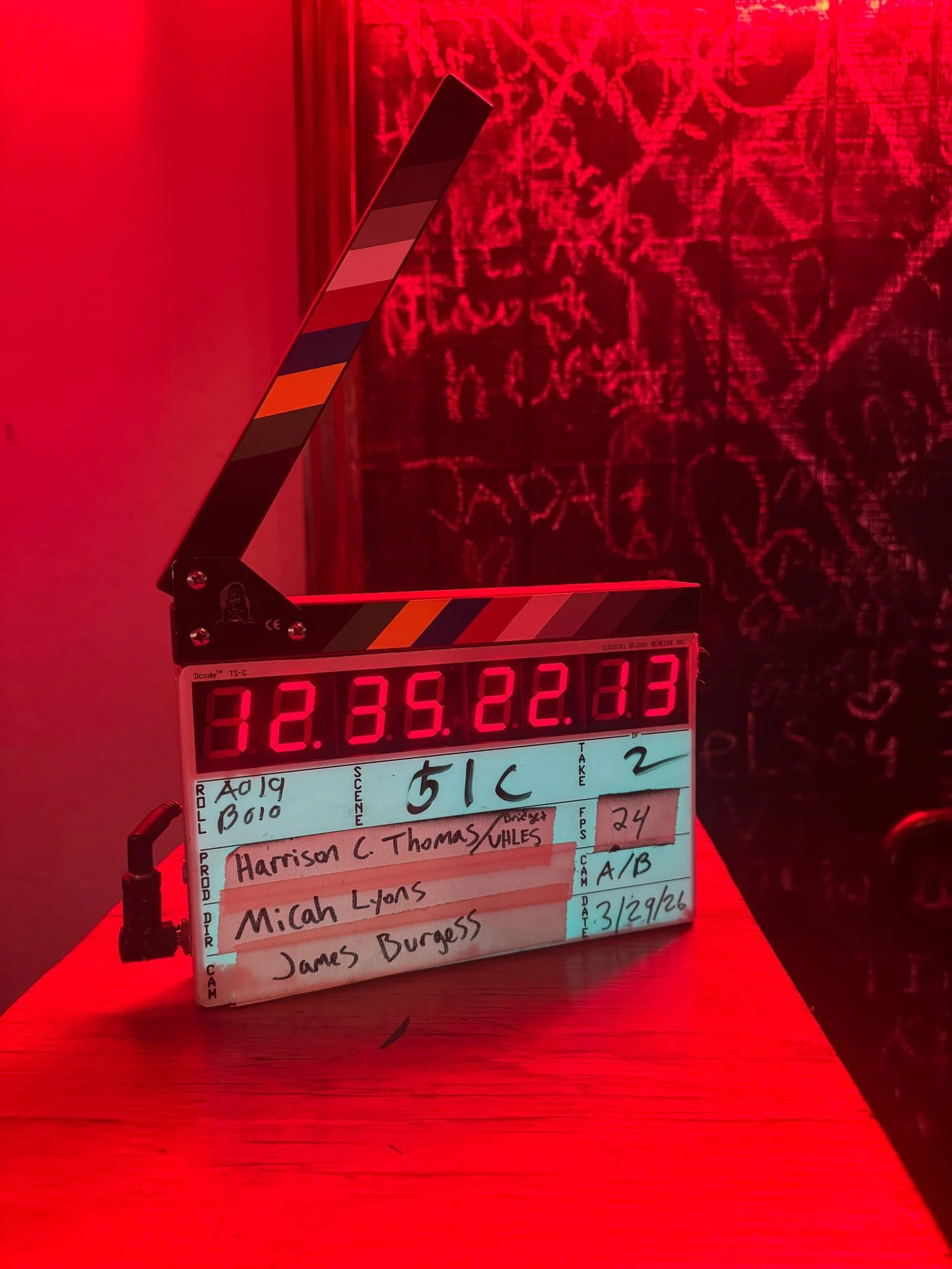 A movie clapboard with a digital timer on top, displaying 12:35:22.13. Beneath the clapboard is a white board with handwritten notes, including names such as Harrison C. Thomas, Micah Lyons, and James Burgess, along with numbers and abbreviations, resting on a wooden table with a red background.