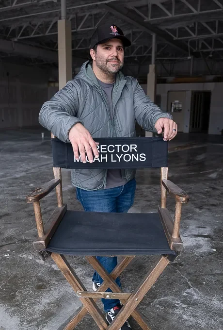 A man stands behind a director's chair with the name Micah Lyons on it, inside a warehouse or industrial space with exposed beams and concrete floor.