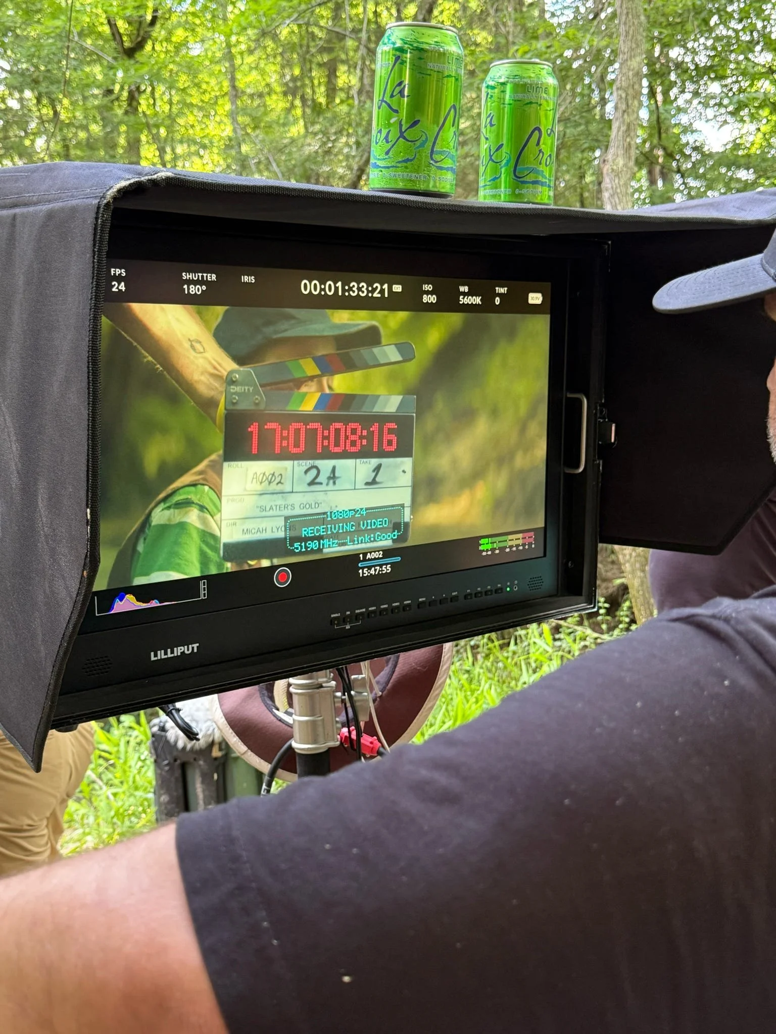 Camera monitor showing a scene with a person in a green shirt holding a clapperboard marked 'Slater's Gold'. Multiple green cans of LaCroix Lime sparkling water are placed on a surface above the monitor, with a forested outdoor background.