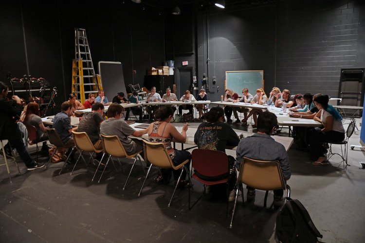 A group of people seated around a large U-shaped table in a dark room, possibly a rehearsal or meeting space, with some equipment and a chalkboard in the background.