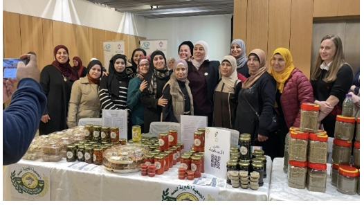 A group of women standing behind a table displaying jars of preserves and packaged food at an indoor market or fair.