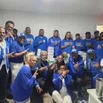 Fraternity brothers wearing blue jerseys and hats, smiling and waving, at a community service event.