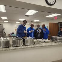 Fraternity brothers posing for a photo in a kitchen, preparing food at a counter at Oak City Cares event in Raleigh, NC.