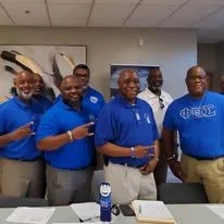 Fraternity brothers in blue polos and brown slacks posing for a photo in a school classroom after a community service event at a local school.