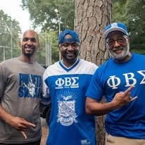 Fraternity brothers standing outdoors posing for a picture at a large tree during fraternity cookout.