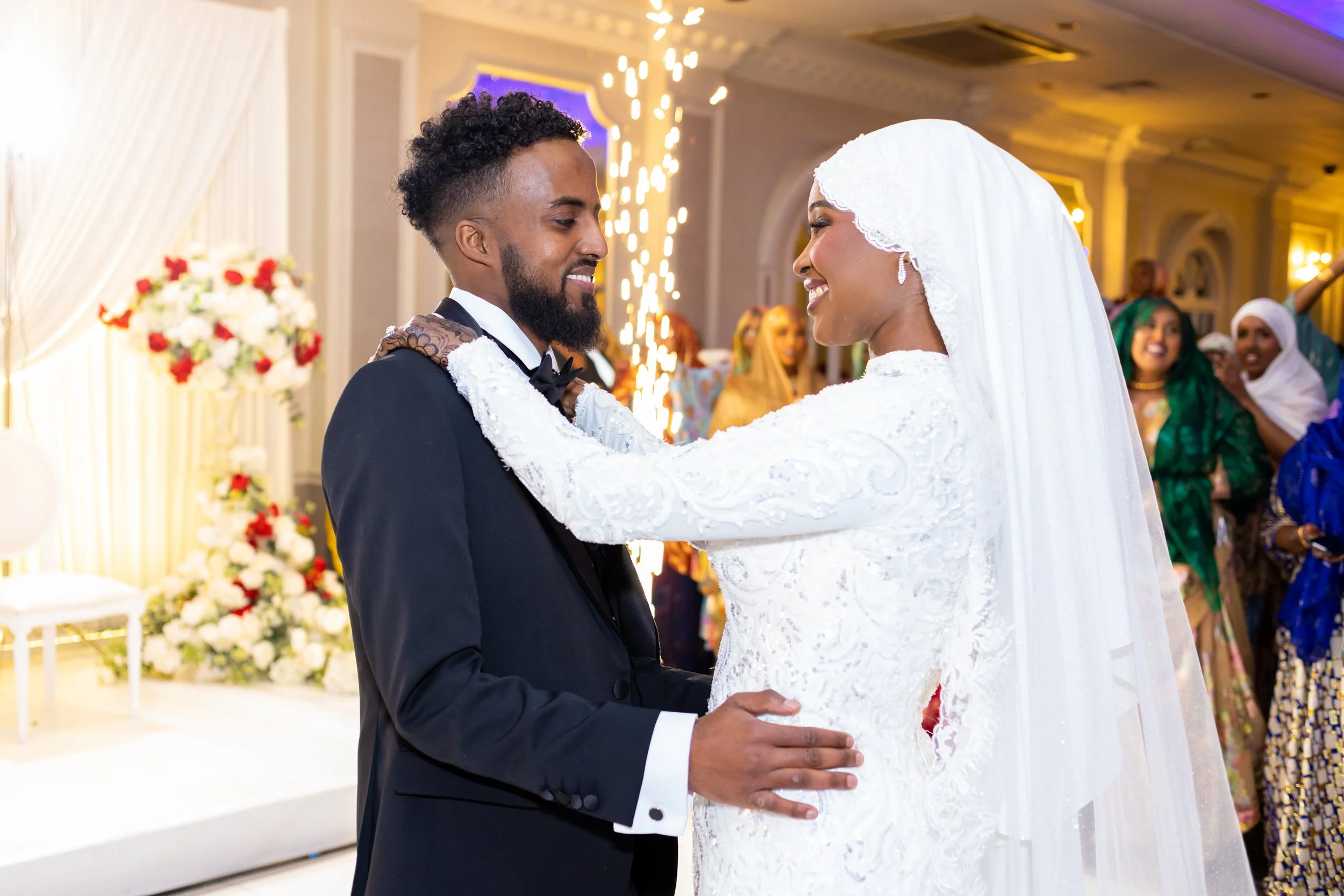 A couple holding hands and looking at each other, dressed in wedding attire, at a wedding reception with friends and floral decorations in the background.