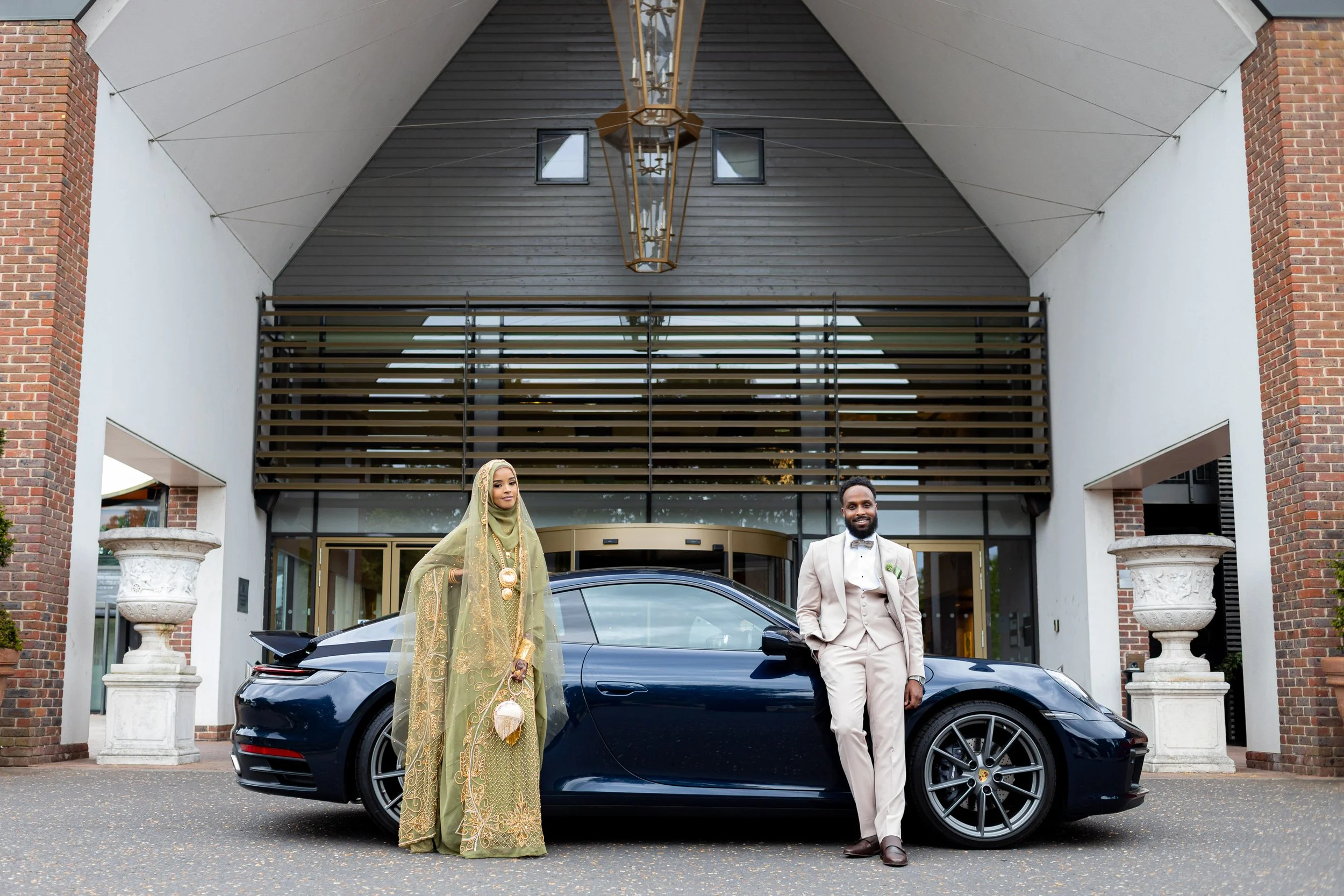 A man and woman dressed in formal attire standing in front of a black luxury car, with a modern building in the background.