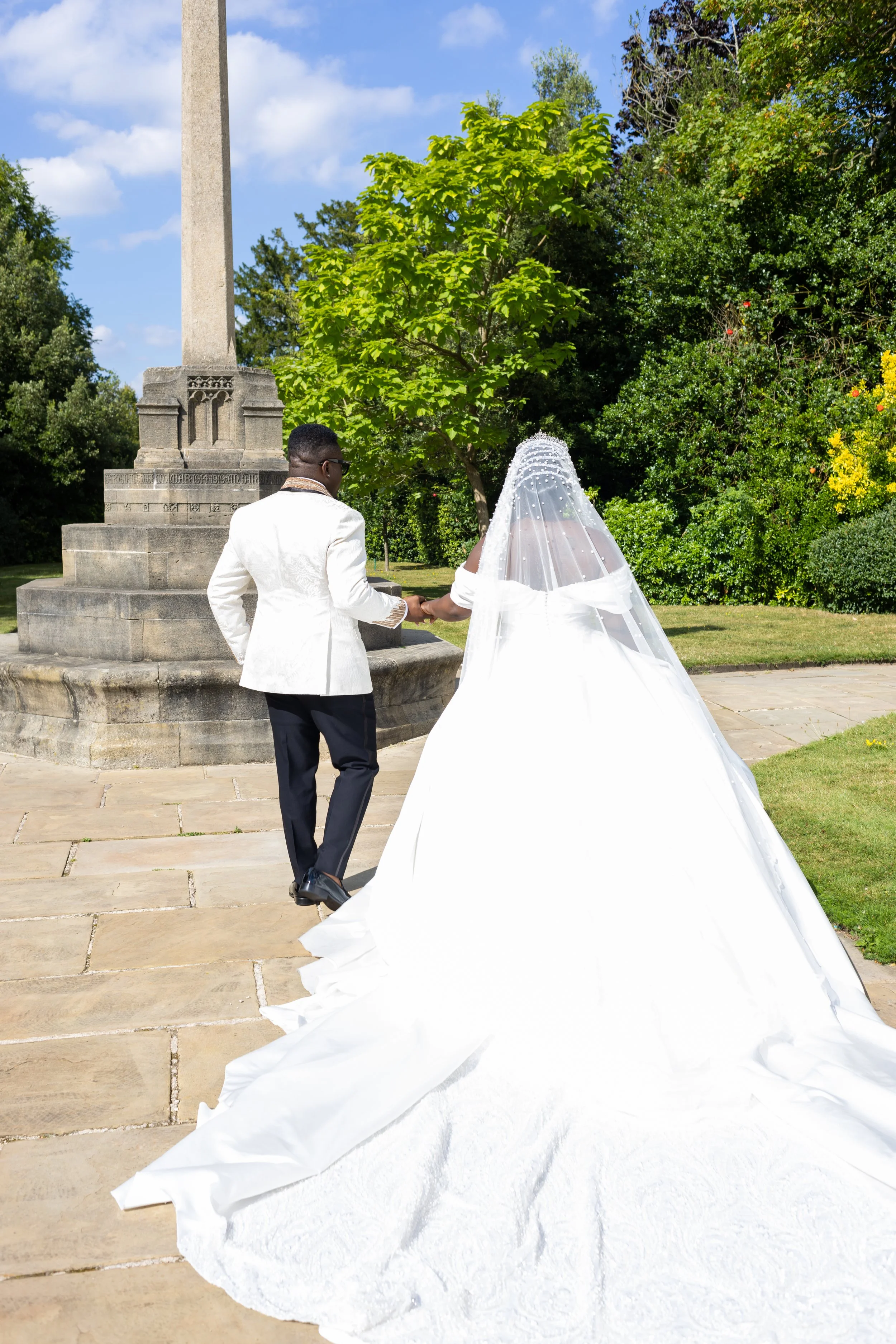 A couple dressed in wedding attire holding hands outdoors near a stone monument, with green trees and a blue sky in the background.