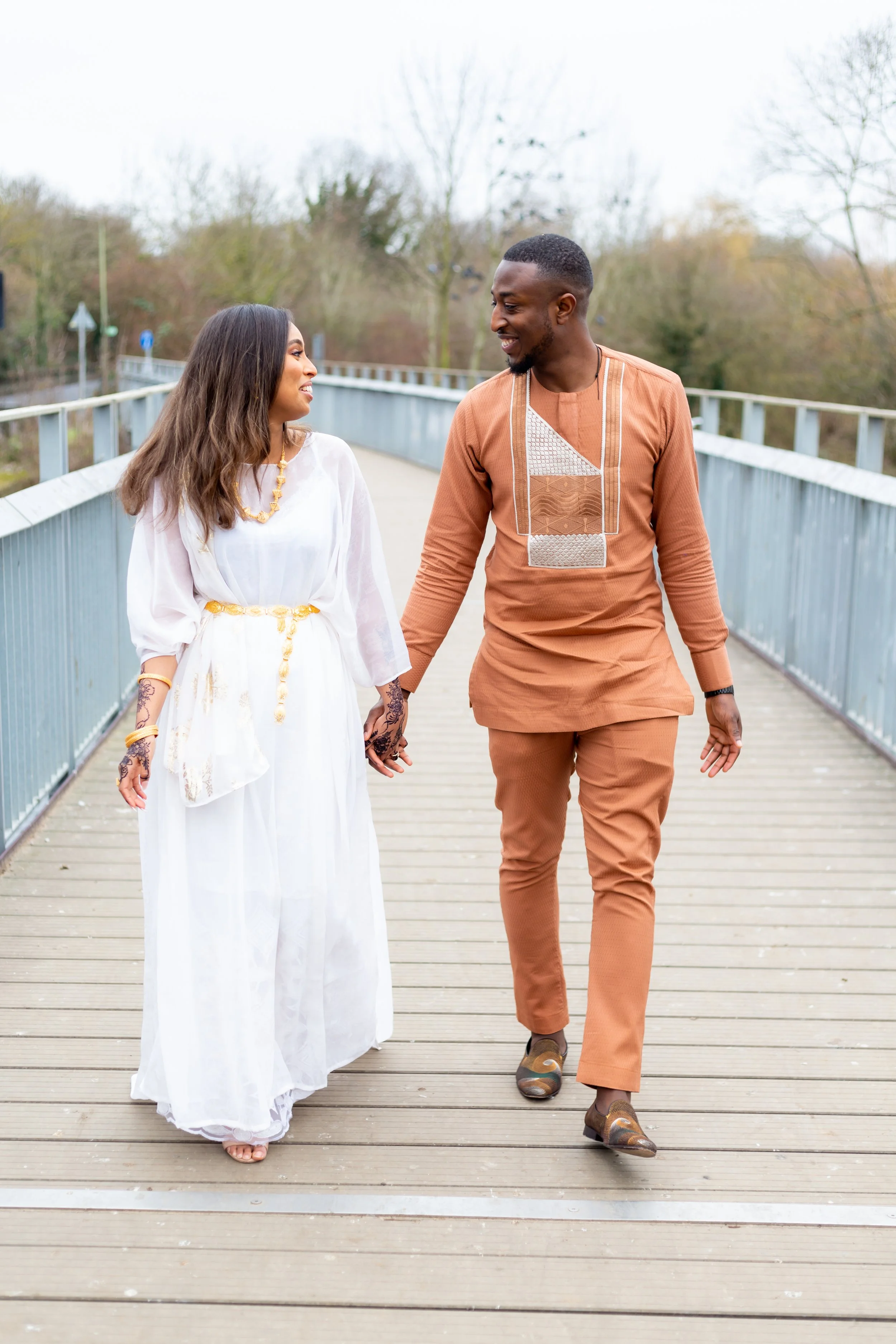 A couple holding hands and walking on a bridge outdoors during the daytime. The woman is wearing a flowing white dress with jewelry, and the man is dressed in a brown traditional outfit. They are smiling at each other, and the background features tre
