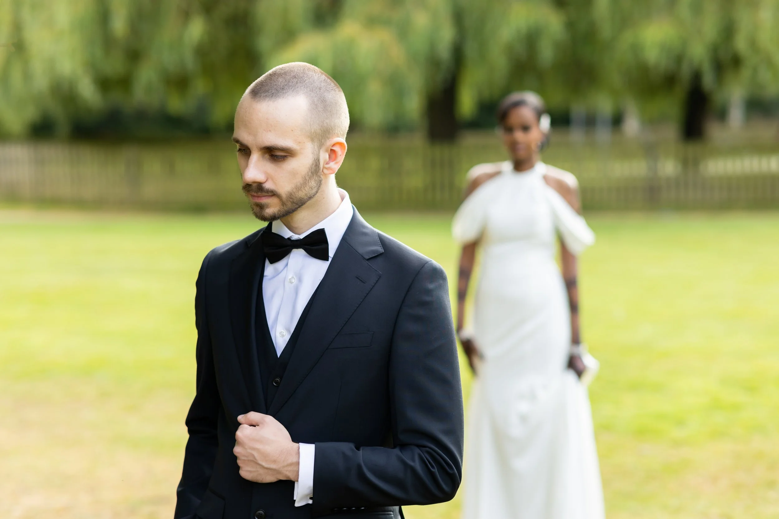 A man in a black tuxedo with a bow tie standing outdoors in the foreground, and a woman in a white dress with a floral pattern and headphones in the background, with trees and a fence in a park setting.