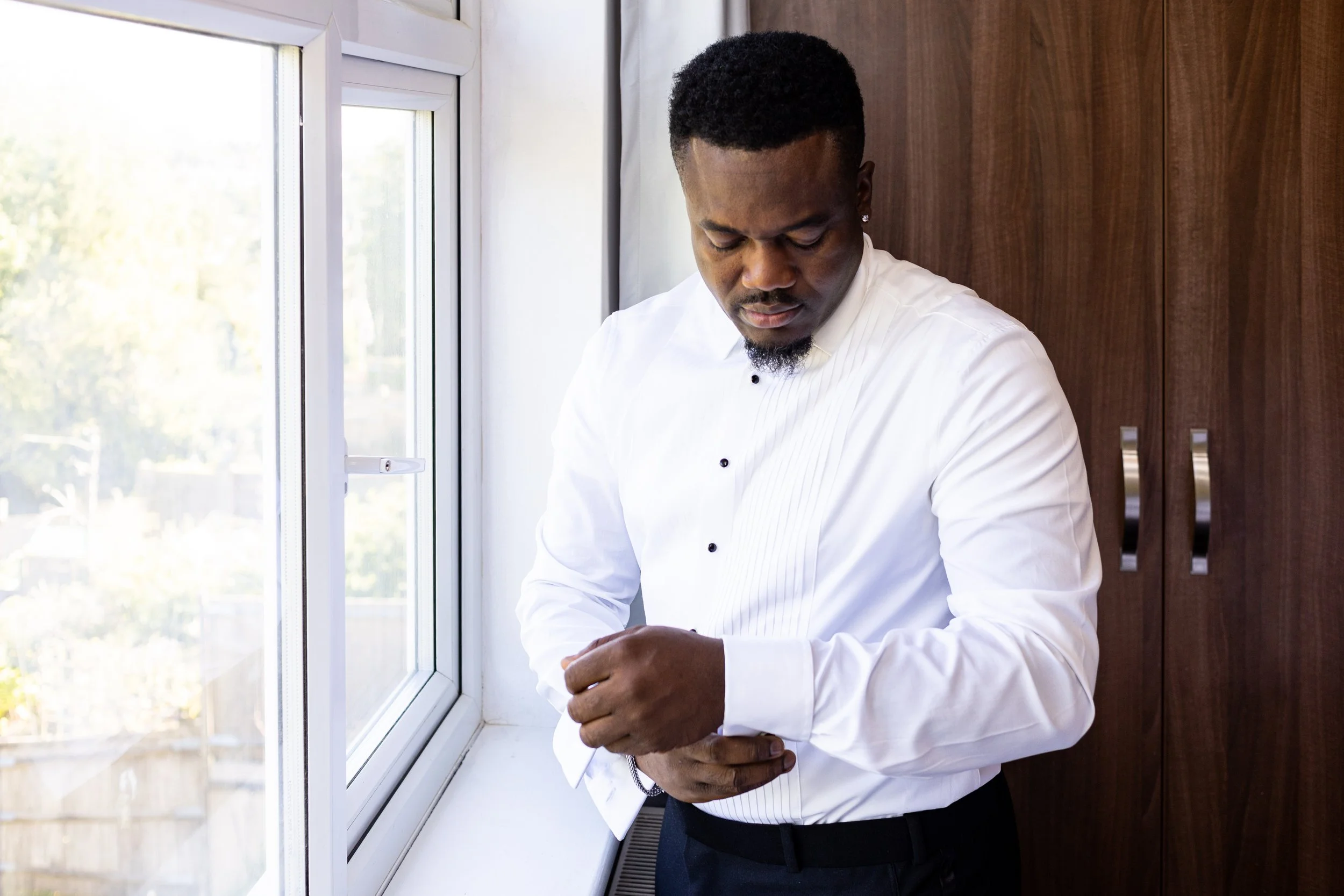 A man in a white shirt adjusts his sleeve near a window inside a room with wooden cabinets.