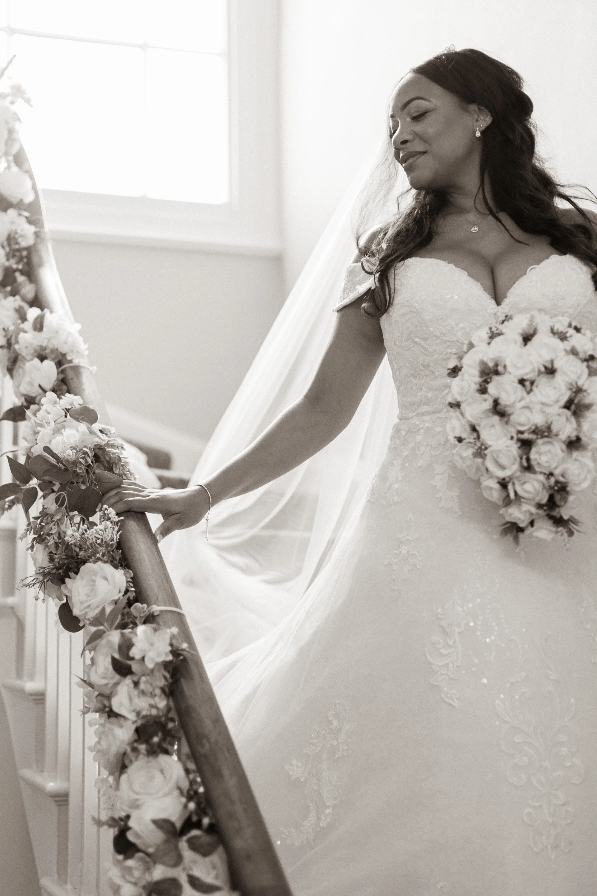 Black and white photo of a bride in a wedding gown holding a bouquet, standing on a staircase with floral decorations.