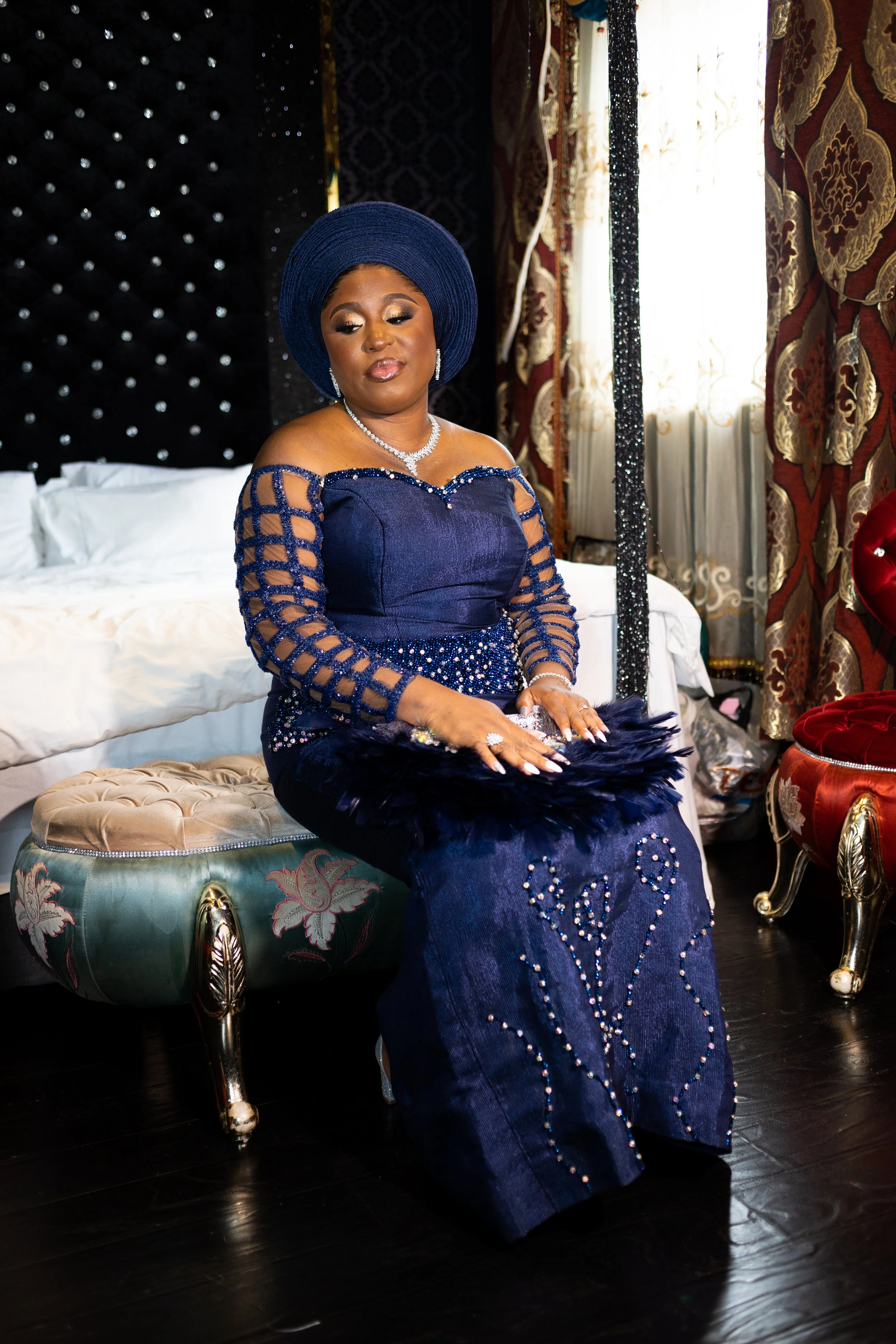 A woman dressed in a dark blue, off-the-shoulder dress with intricate beading sits on a tufted stool. She wears a matching blue head wrap and elegant jewelry, including earrings and a necklace. The background features patterned curtains with gold and