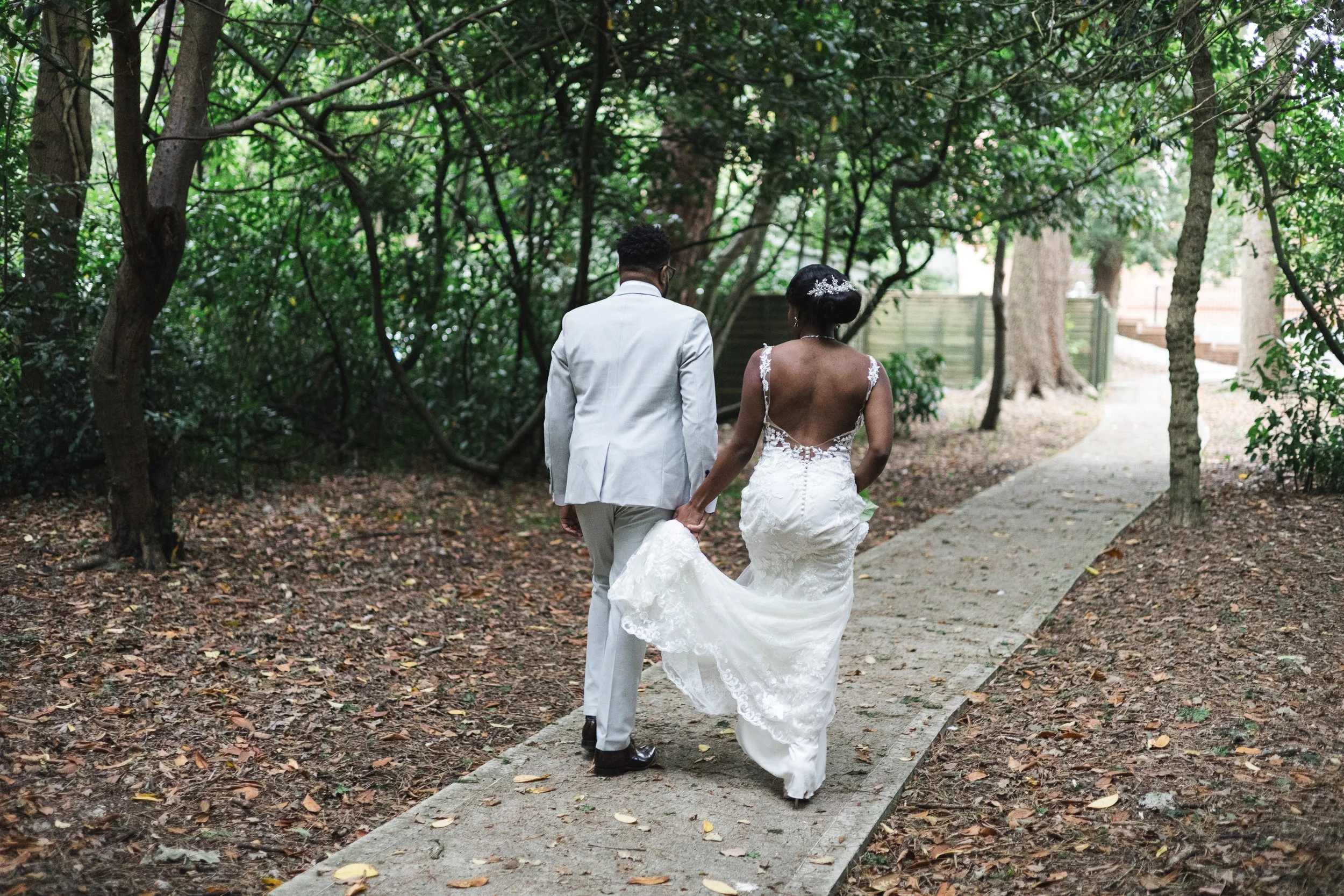 A newlywed couple walking hand in hand through a wooded park, with the bride in a lace wedding gown and the groom in a light-colored suit.